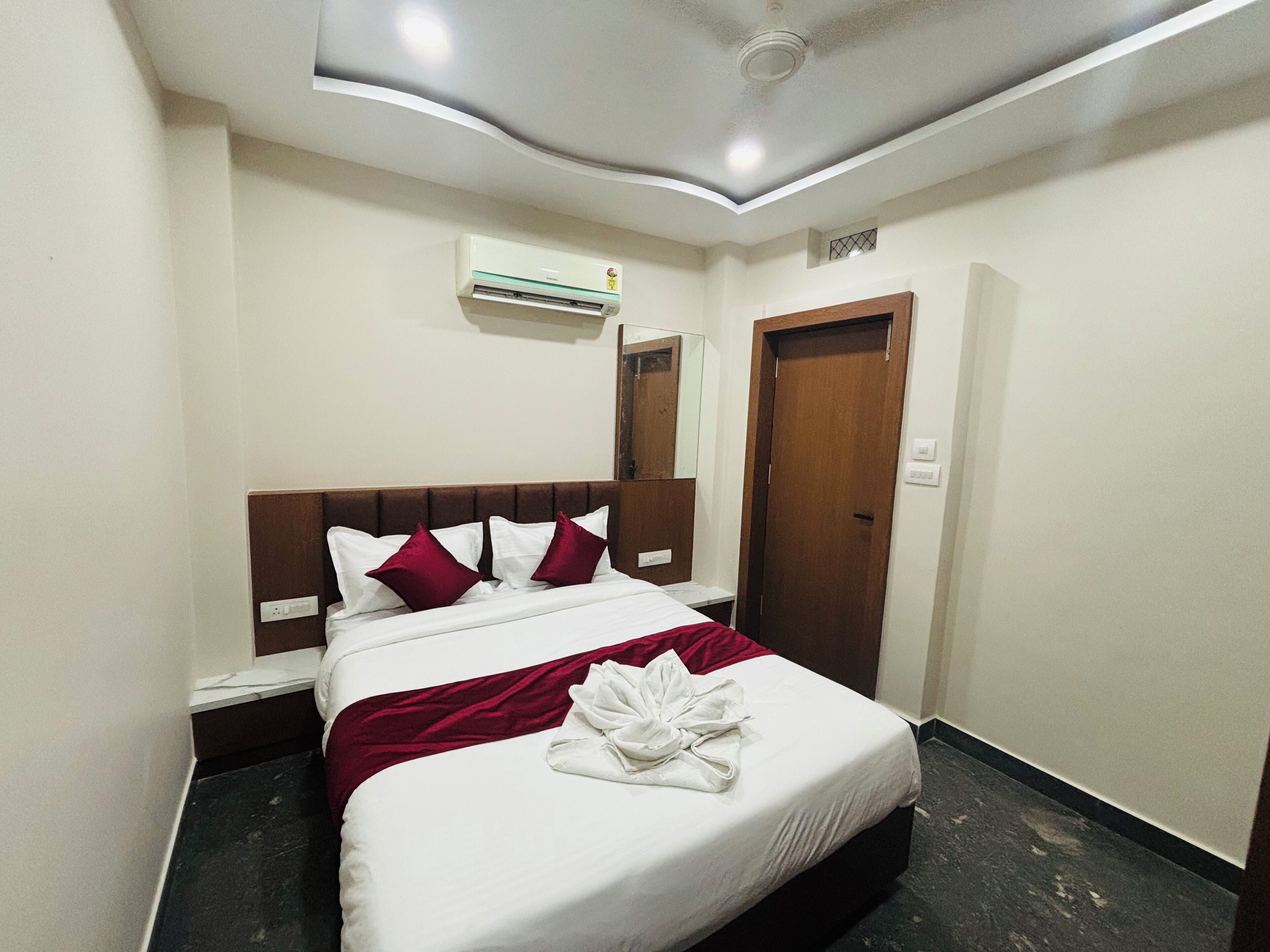 Habitación de hotel con cama, cabecera de madera, aire acondicionado blanco, puerta de madera y espejo en la pared.