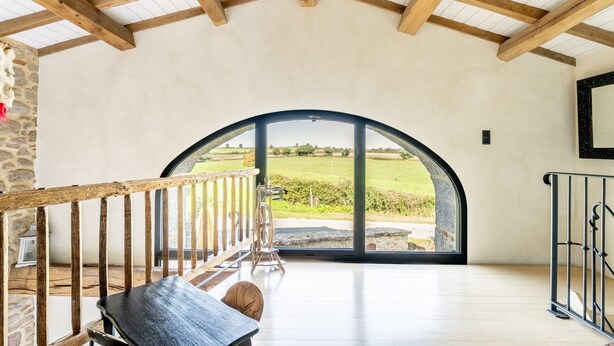 Ein geräumiger Raum mit hoher Decke, Holzbalken, einem großen Rundbogenfenster und Blick auf eine grüne Landschaft.