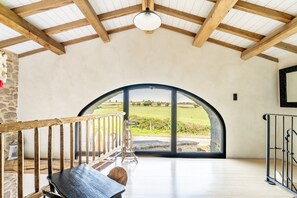 A spacious room with a high ceiling, wooden beams, a large arched window, and a view of a green landscape.