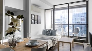 Una habitación de hotel moderna con un ventanal grande, una cama con vistas, una zona de comedor con una mesa para dos, y un jarrón con flores.