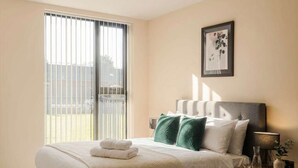 Une chambre à coucher avec un lit, une table de chevet, une lampe, un tableau encadré et une fenêtre avec des stores.