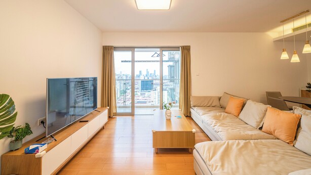 Ein modernes Wohnzimmer mit großem Flachbildfernseher, einem Holztisch, einem beige farbenen Sofa und Blick auf die Stadt durch große Fenster.