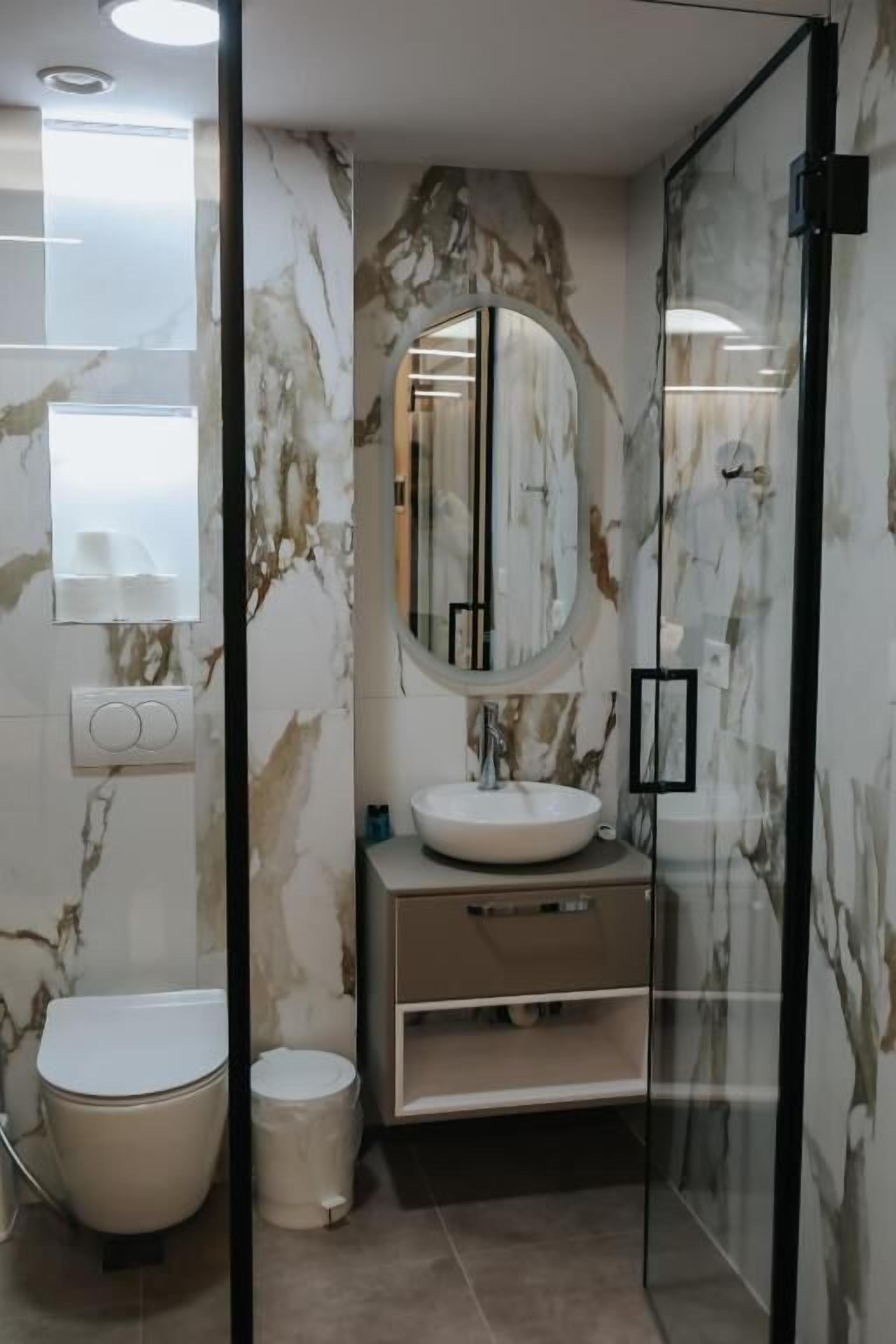 A modern bathroom with a marble wall, a round mirror, a sink, and a toilet.
