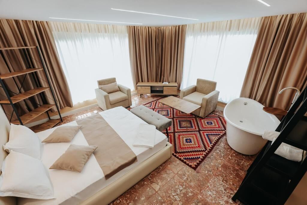 A hotel room with a large bed, two armchairs, a sofa, a small table, a bathtub, and a wooden shelf.