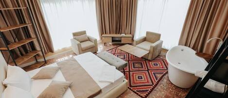 A hotel room with a large bed, two armchairs, a sofa, a small table, a bathtub, and a wooden shelf.