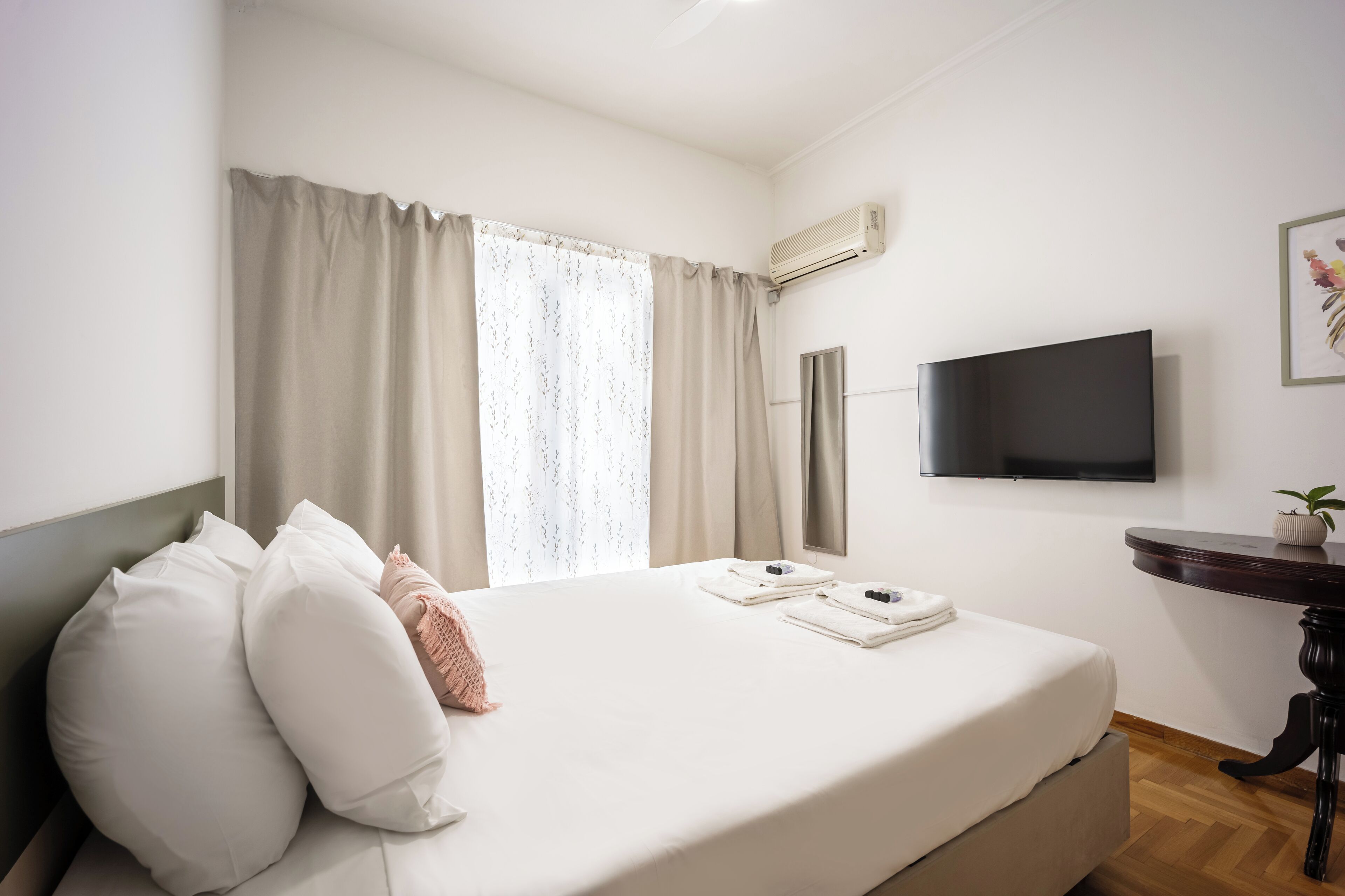 A hotel room with a bed, a flat-screen TV, a small table, and a window with curtains.