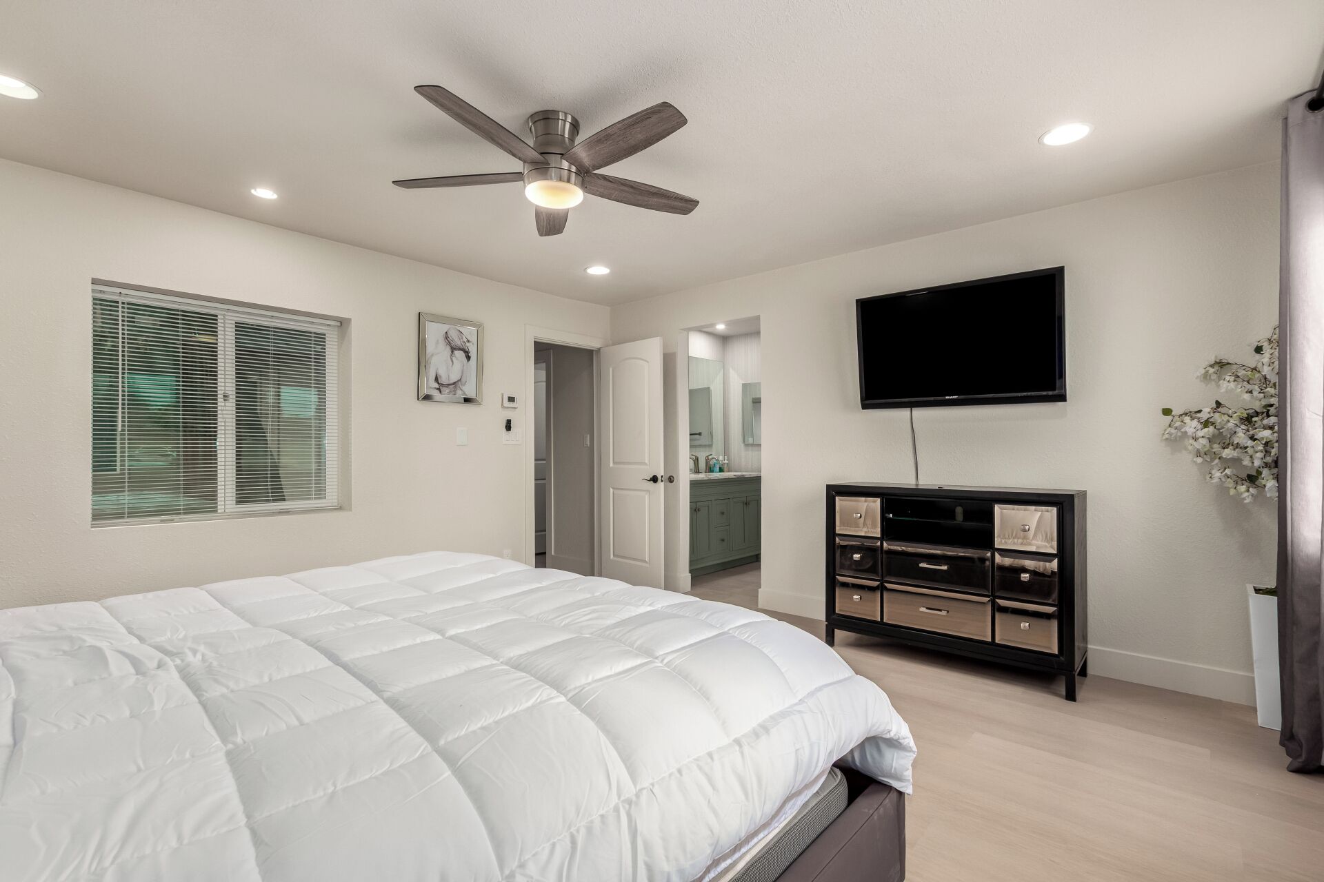 A bedroom with a bed, a ceiling fan, a TV, a dresser, and a bathroom visible through an open door.