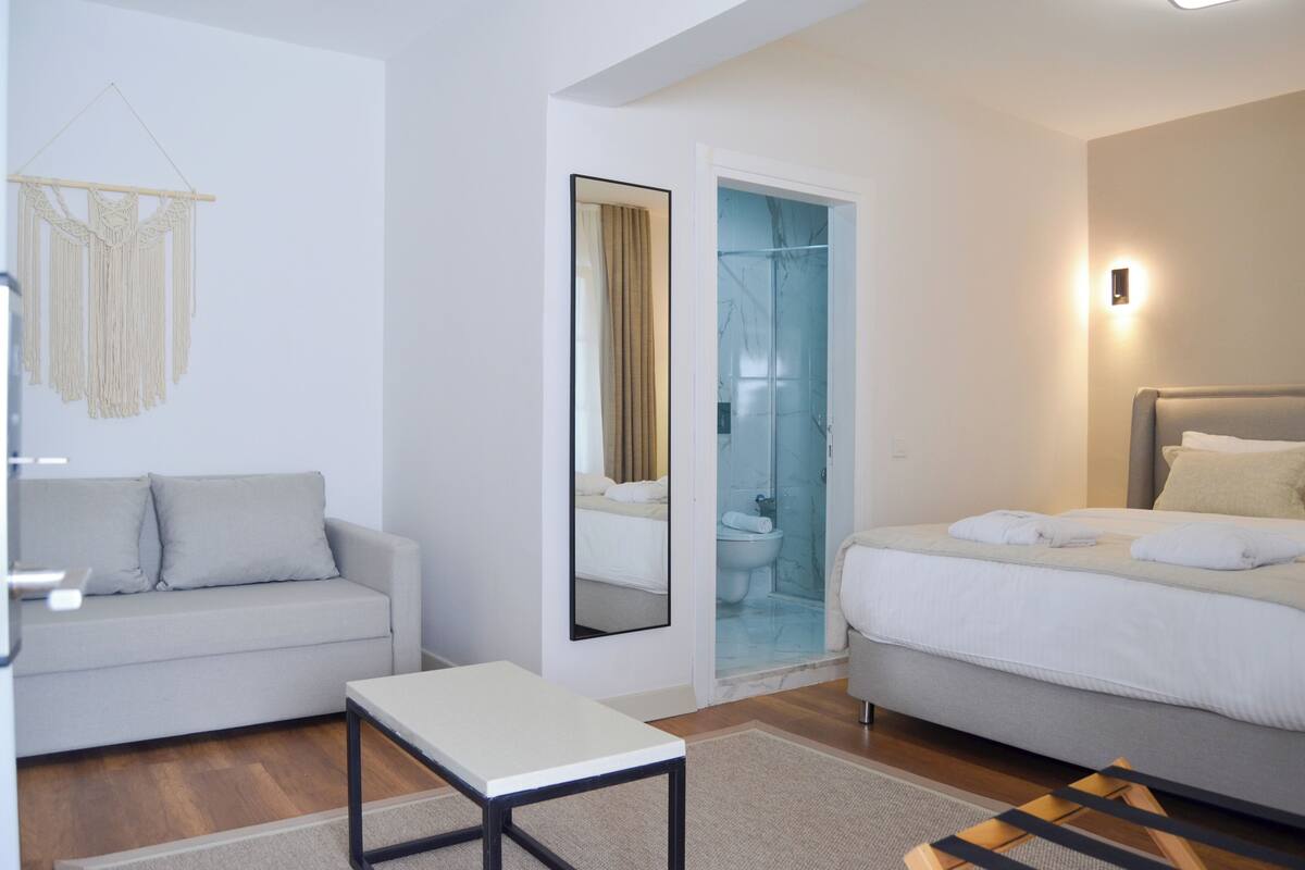 A modern hotel room with a sofa, a bed, a small table, and a glass-enclosed shower area.