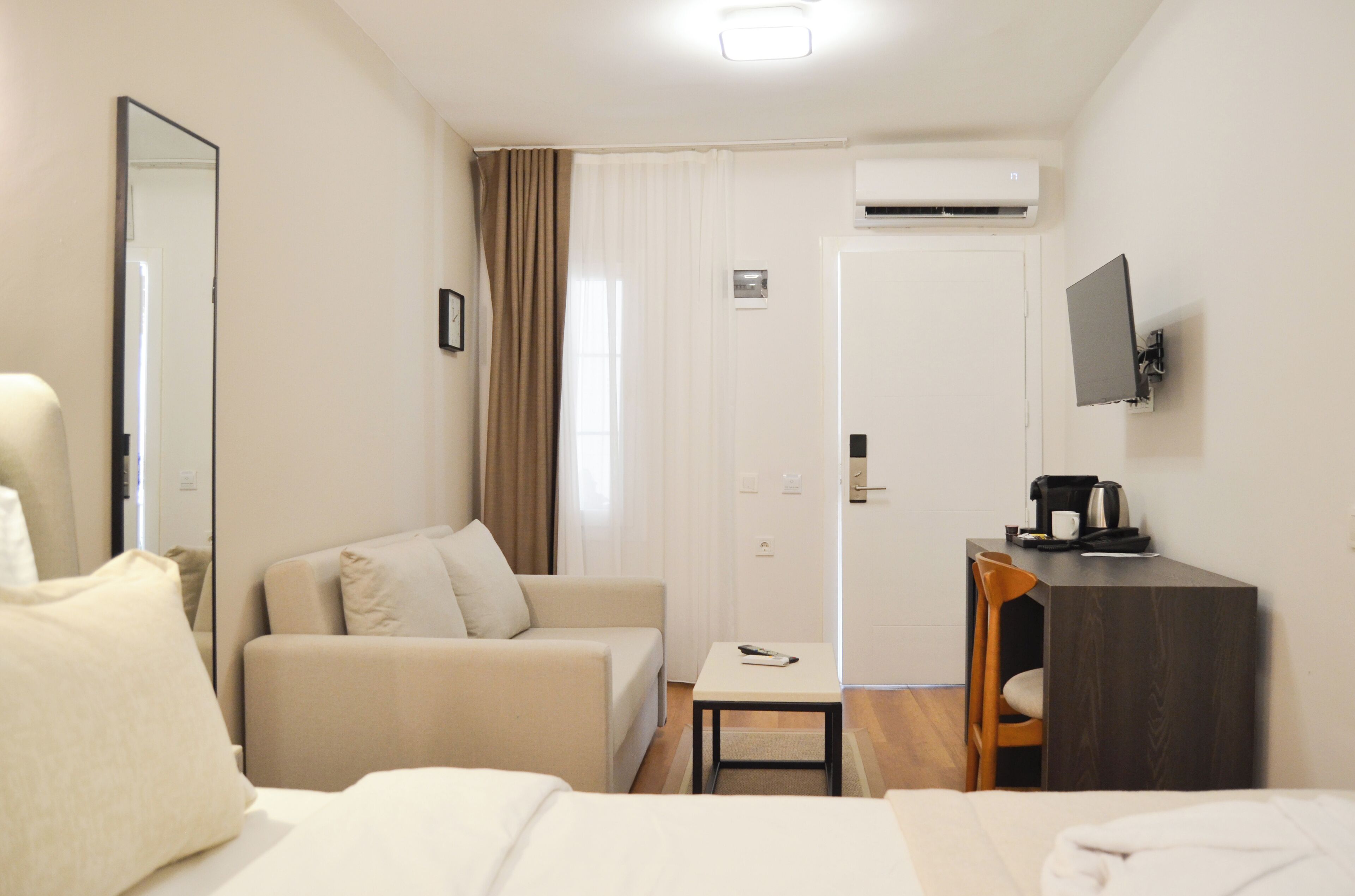 A hotel room with a bed, a sofa, a small table, a chair, a desk with a computer, and an air conditioning unit.