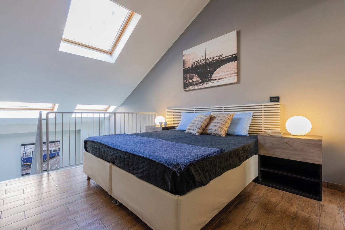 A bedroom with a skylight, a bed with a headboard, bedside lamps, and a painting of a bridge.