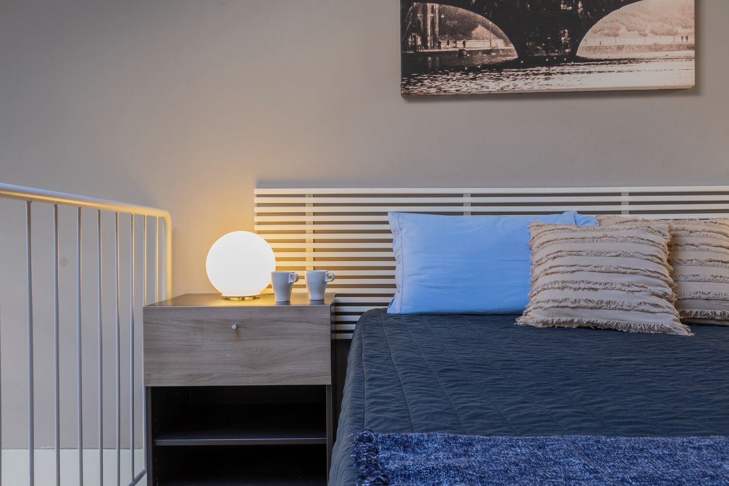 A bedroom with a bed, bedside table, lamp, and a wall-mounted picture.