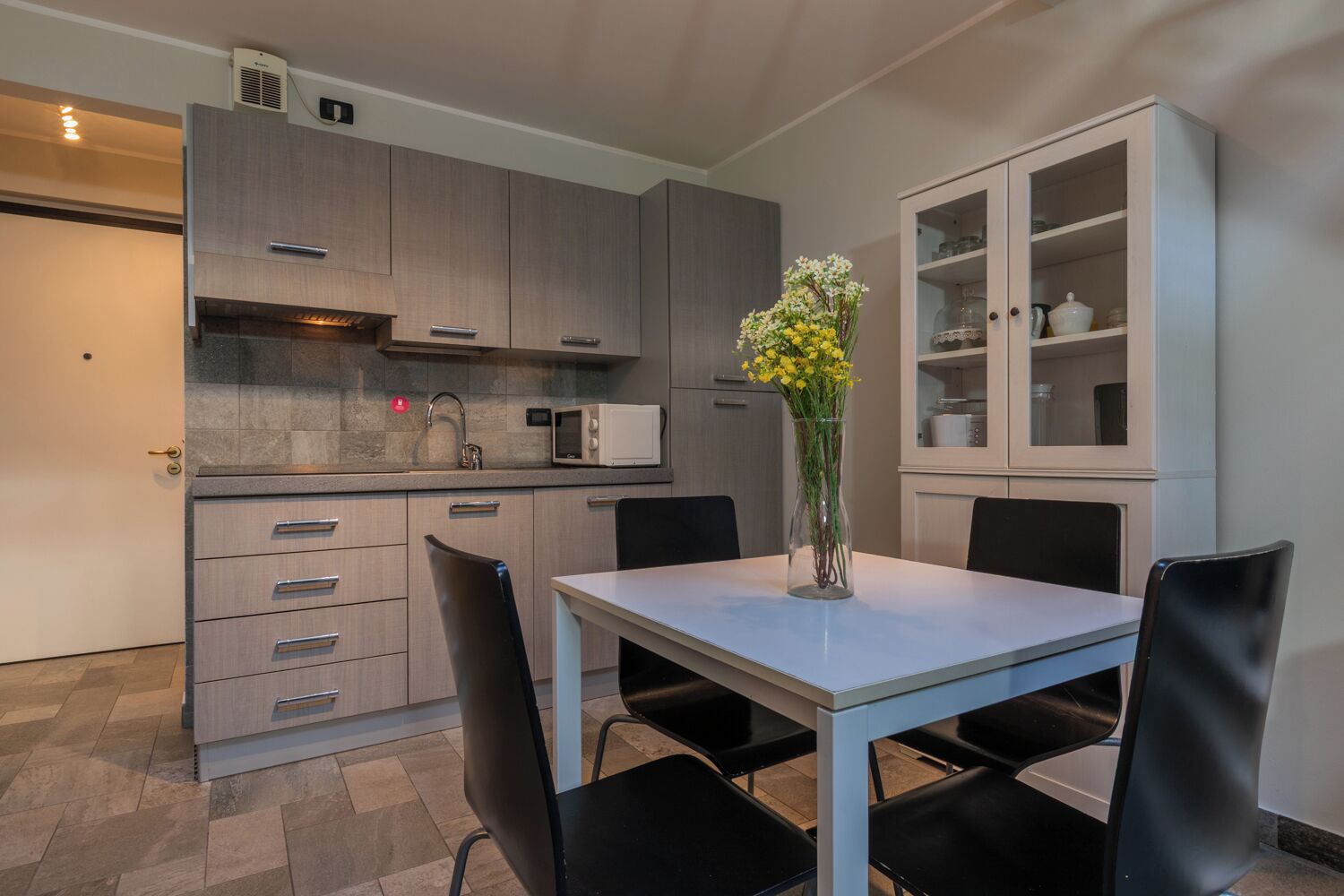Une cuisine moderne avec des meubles gris, un micro-ondes encastré et un coin repas comprenant une table blanche et des chaises noires.