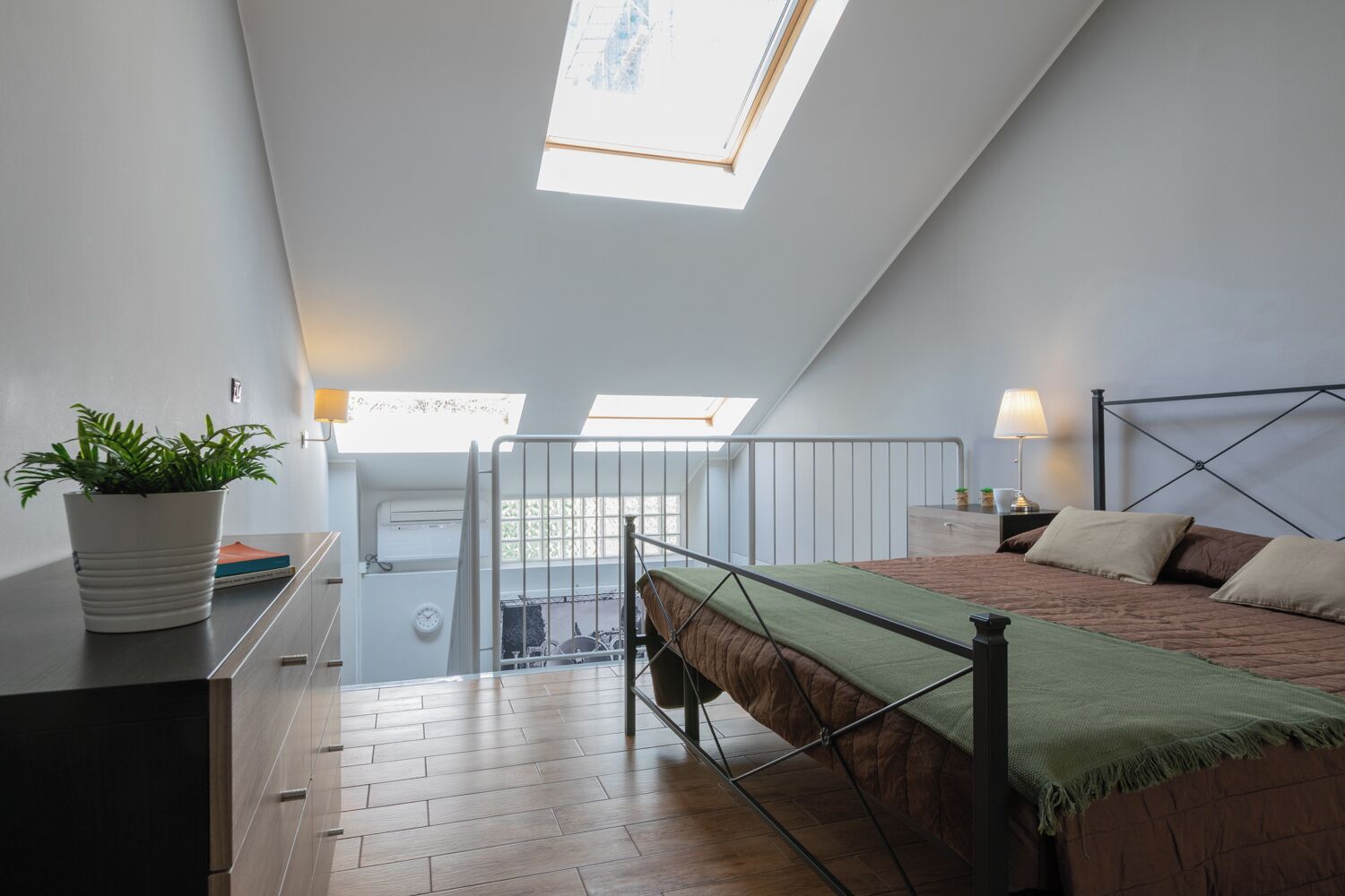 Une chambre avec une lucarne, un lit recouvert d’un plaid vert, une table de chevet avec une lampe et une plante en pot posée sur une commode.