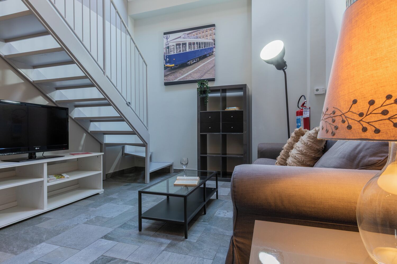Ein modernes Wohnzimmer mit Treppe, Flachbildfernseher, Glastisch, Sofa, Stehlampe und Bücherregal.