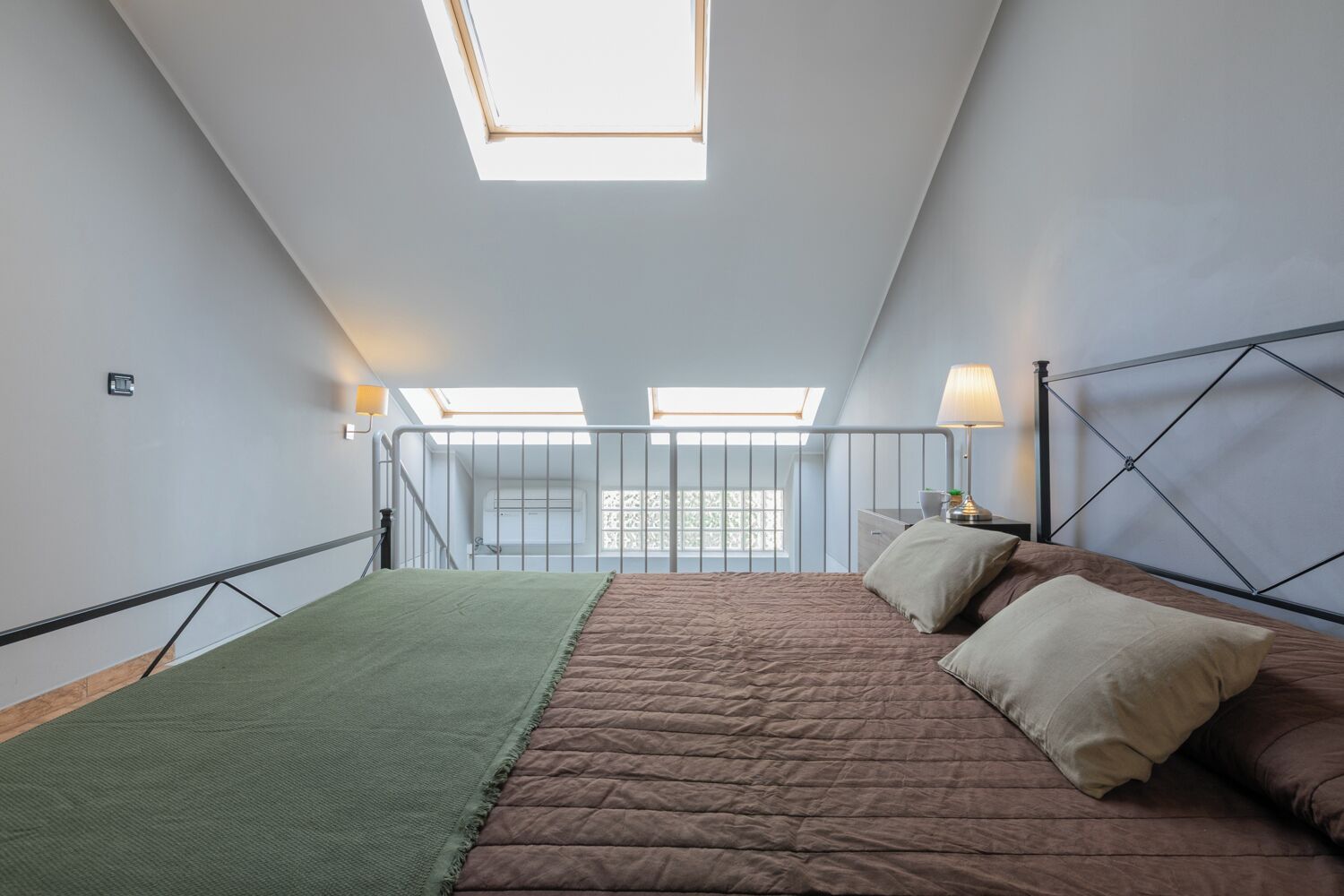Ein Schlafzimmer mit Dachfenster, einem Bett mit brauner Steudecke und einem Nachttisch mit Lampe.