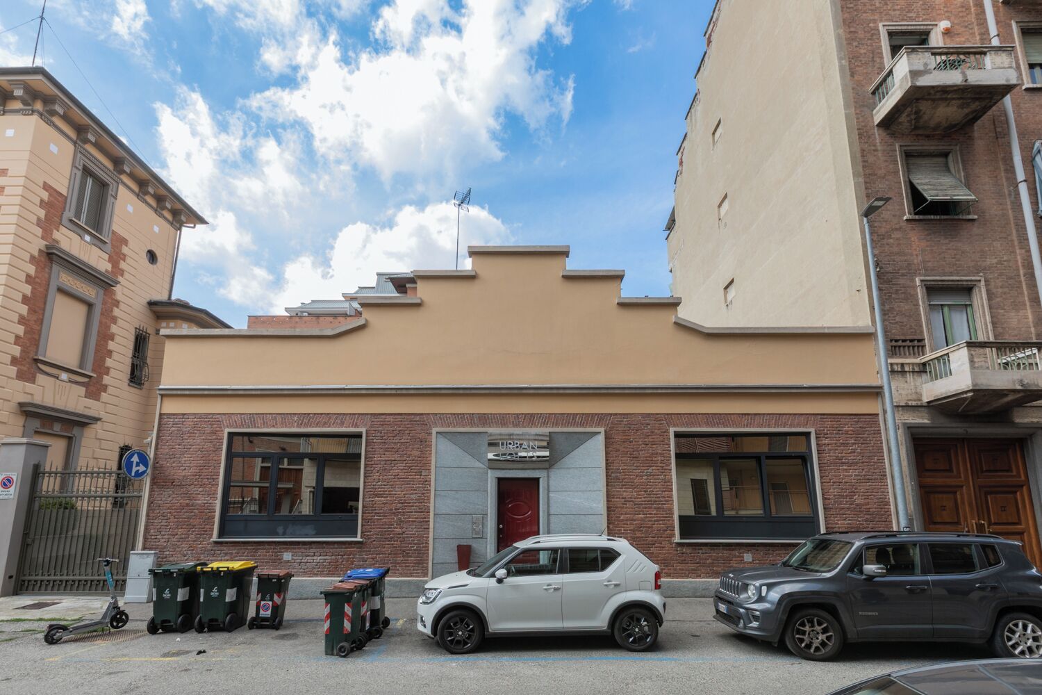 Un edificio con fachada color beige, una puerta roja y dos ventanas. Hay tres coches estacionados adelante.