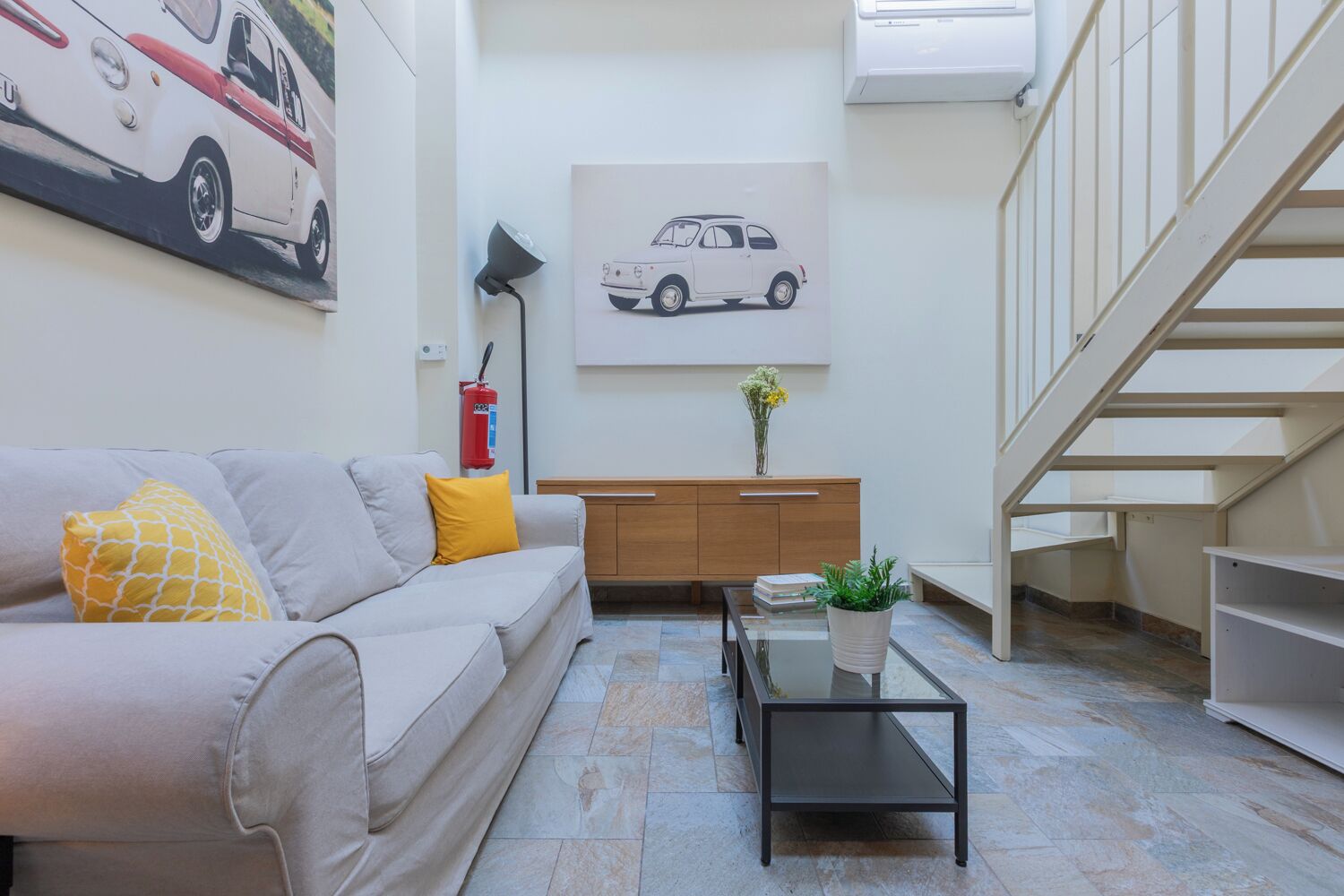 A modern living room with a sofa, a coffee table, a painting, a fire extinguisher, and a staircase.