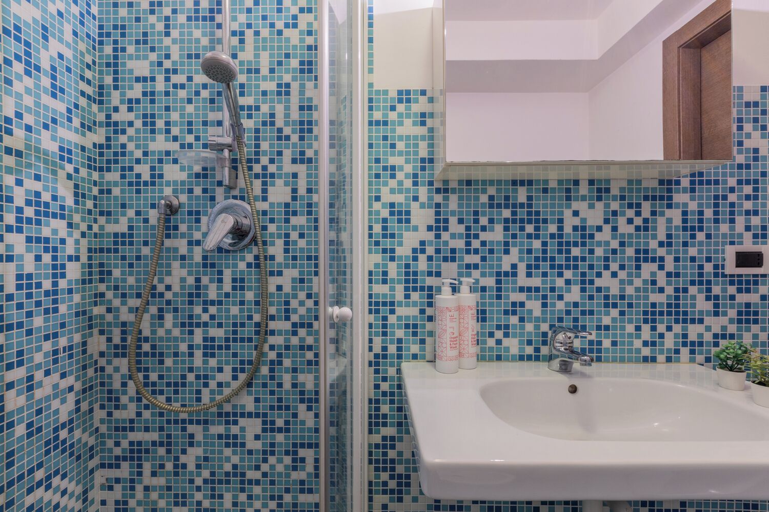 Casa de banho com chuveiro e lavatório, revestida com mosaicos azuis e brancos.