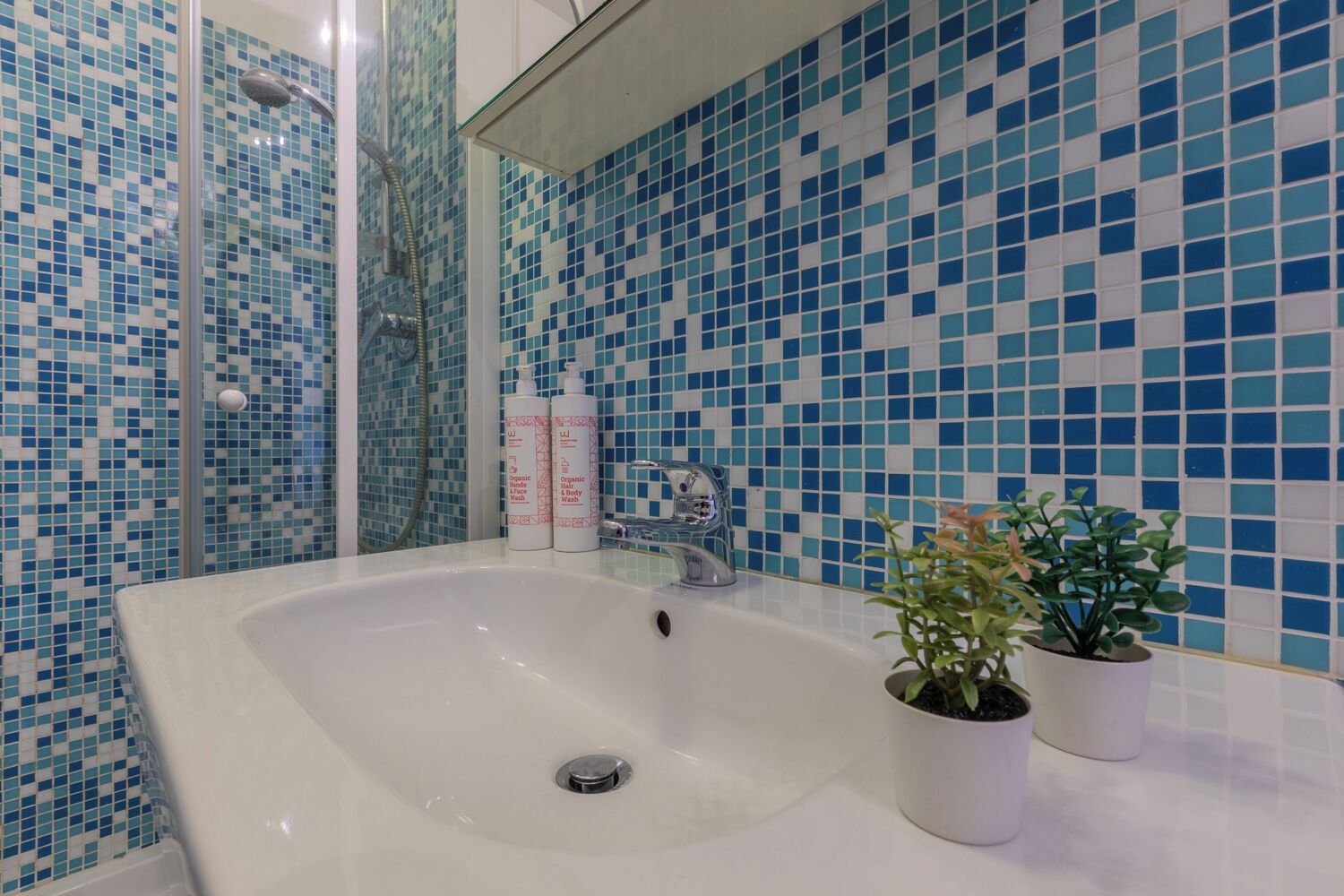 A bathroom with a large white sink, two potted plants, and a shower with blue mosaic tiles.