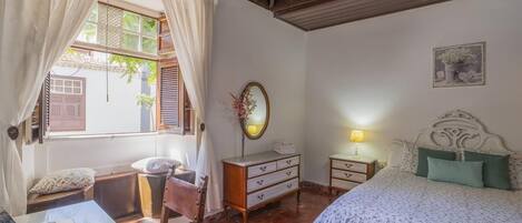 A bedroom with a bed, a desk with a laptop, a chair, a dresser, and a bedside table with a lamp.