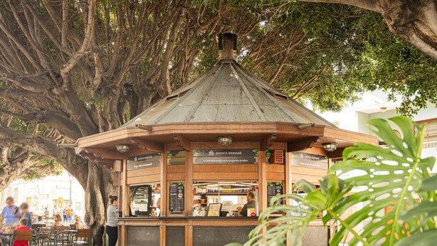 A gazebo-style kiosk with a wooden roof and glass front, surrounded by outdoor seating and greenery.