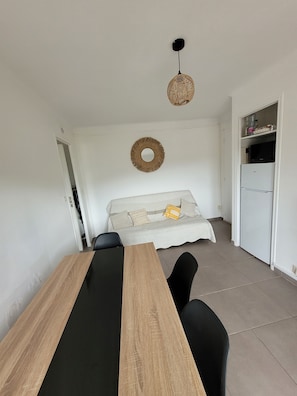 A dining area with a wooden table and chairs, a sofa with cushions, a hanging light fixture, and a refrigerator. - One-bedroom flat sleeps 4 seaside location, residence in Fabrégas, Les Sablettes (La Seyne-sur-Mer)