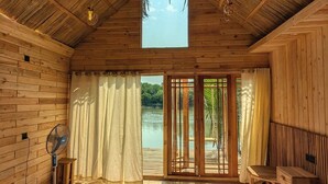 Quarto de cabana de madeira com cama, ventilador e vista para um lago através de uma porta de vidro.
