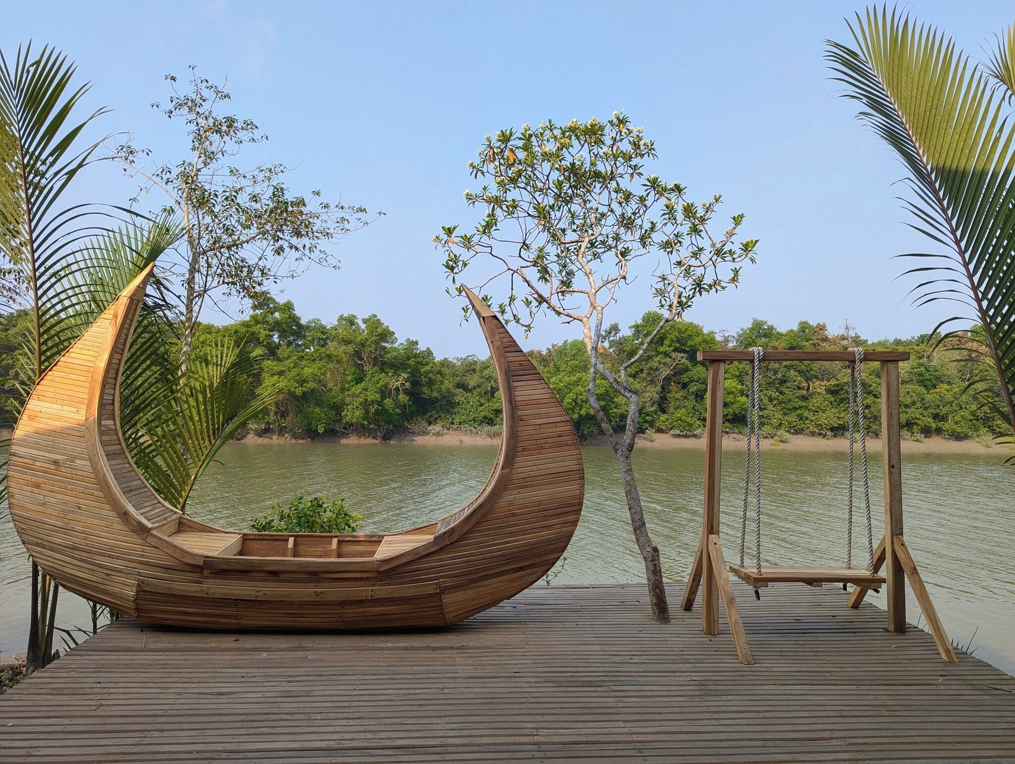 A wooden boat-shaped seating area on a wooden deck by a lake, with a swing set and trees in the background.