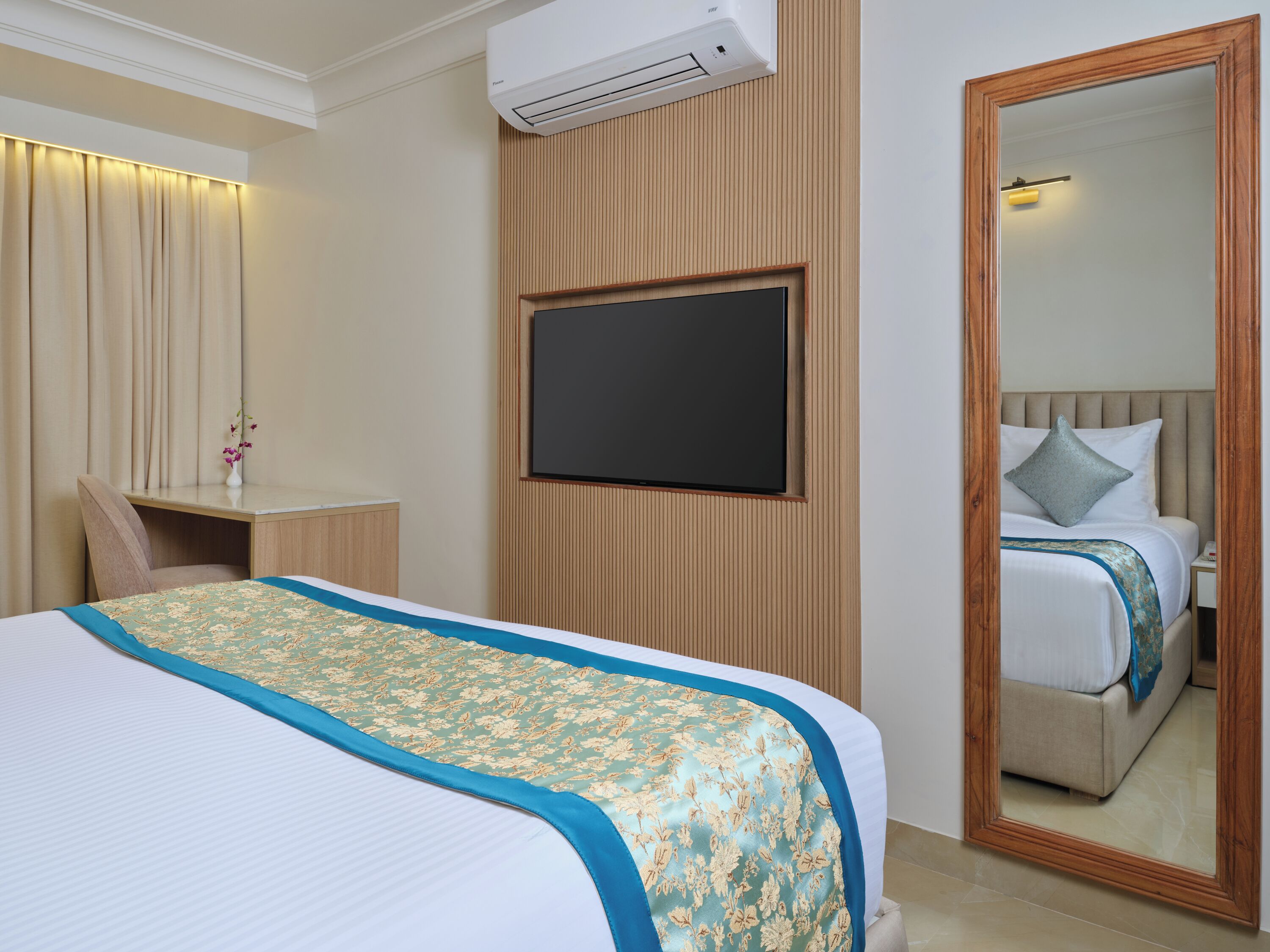 A hotel room with a bed, a desk, a chair, a TV, an air conditioner, and a mirror.