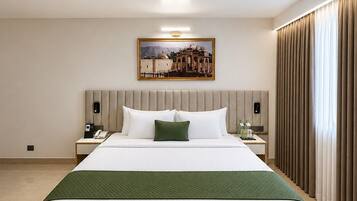 A hotel room with a large bed, a nightstand with a phone, a painting on the wall, and a window with curtains.