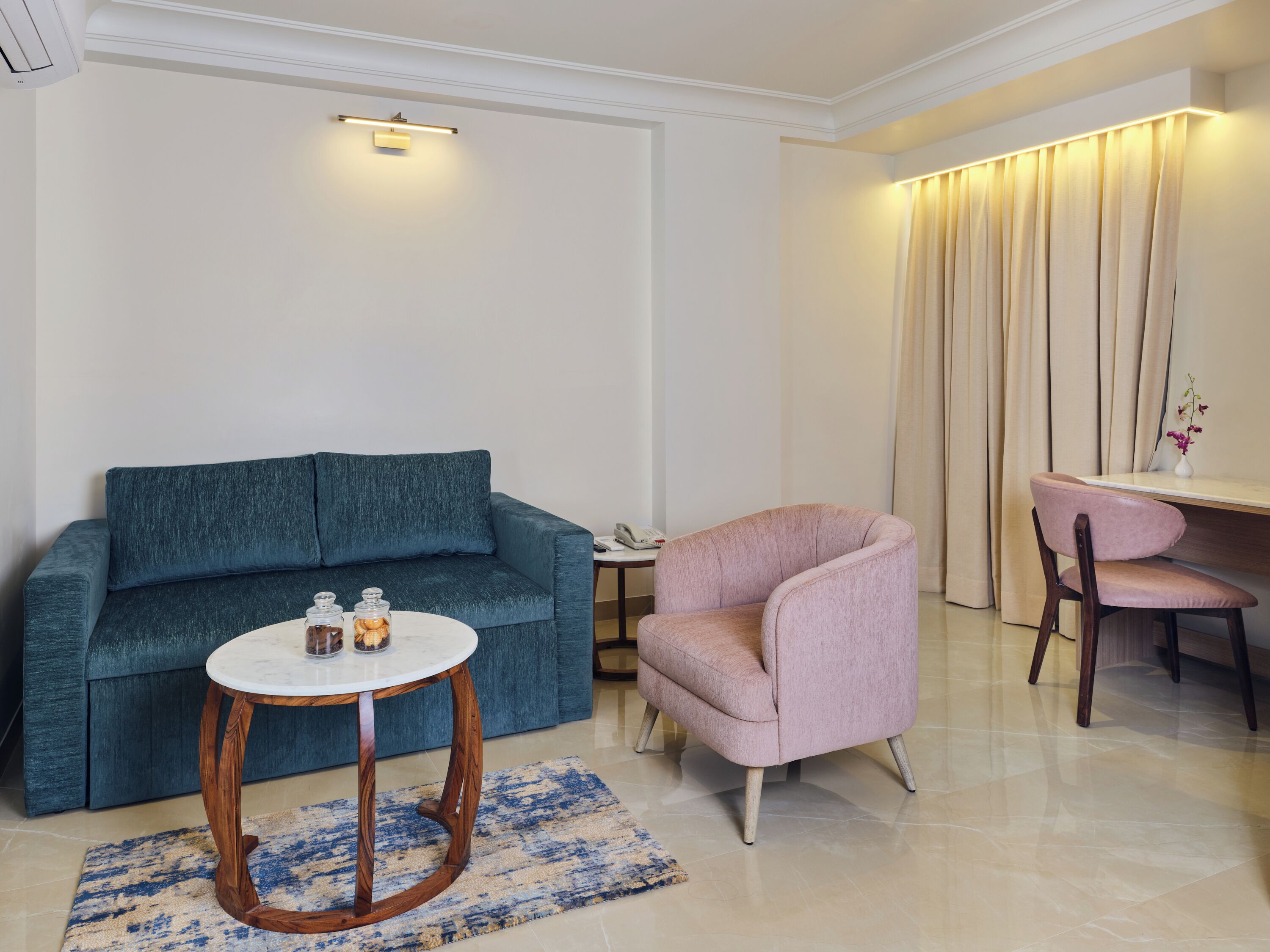 Una sala de estar con un sofá azul, una mesa redonda de madera, un sillón rosa y un escritorio con una silla.