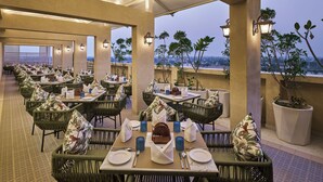 A rooftop dining area with tables set for two, surrounded by wicker chairs, under a covered structure with a view of the cityscape. - Regenta Anantam Resort Khatu Shyam Ji  (Danta Ramgarh)