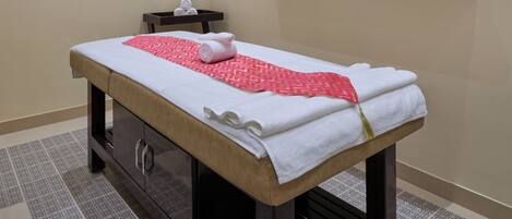 A massage table with towels and a pink patterned cloth, wooden shelves with decorative items, and a small statue of a person in a meditative pose.