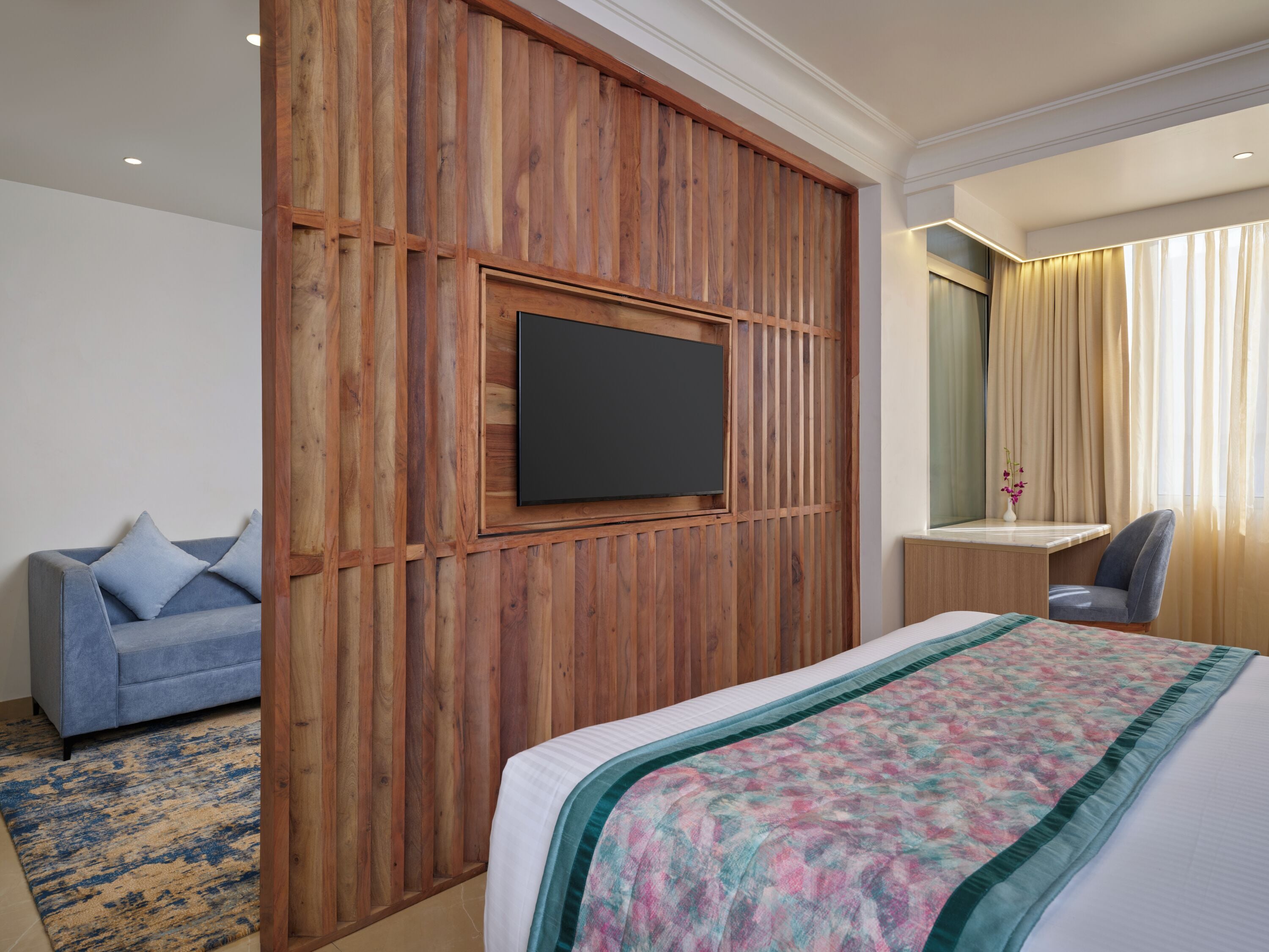 Ein Hotelzimmer mit Holzpaneelen, einem Flachbildfernseher, einem Bett mit floralem Überwurf, einem blauen Sofa, einem Schreibtisch mit Stuhl und einem Fenster mit Vorhängen.