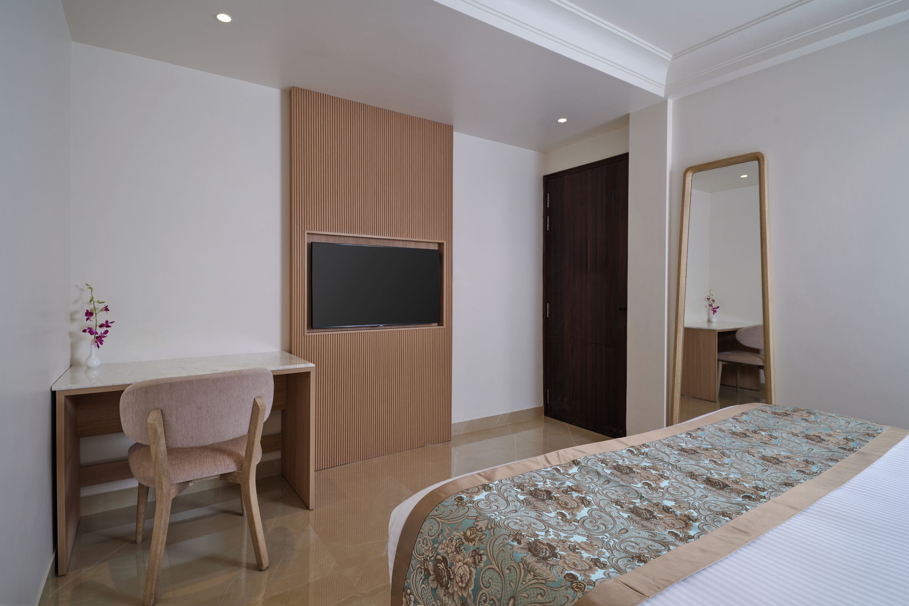 A bedroom with a bed, a desk, a chair, a TV, a mirror, and a wardrobe.