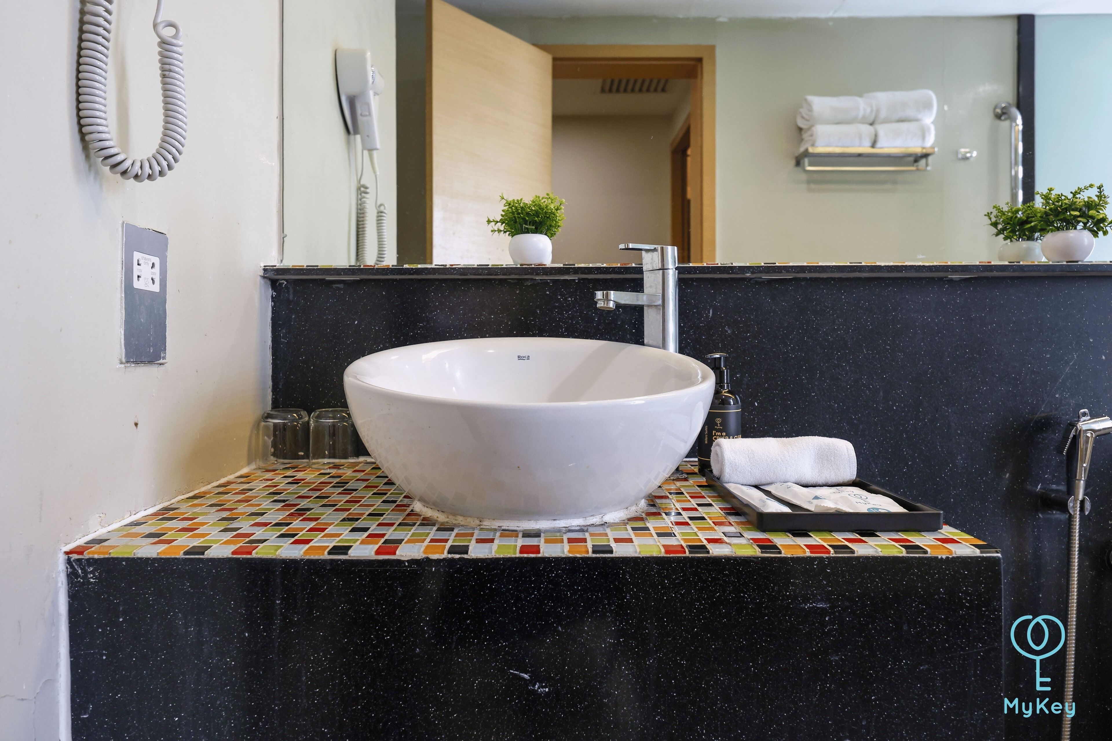 Un baño con un lavamanos blanco de diseño, una encimera negra con un estampado de azulejos coloridos, un espejo, un perchero y una pequeña planta en maceta.