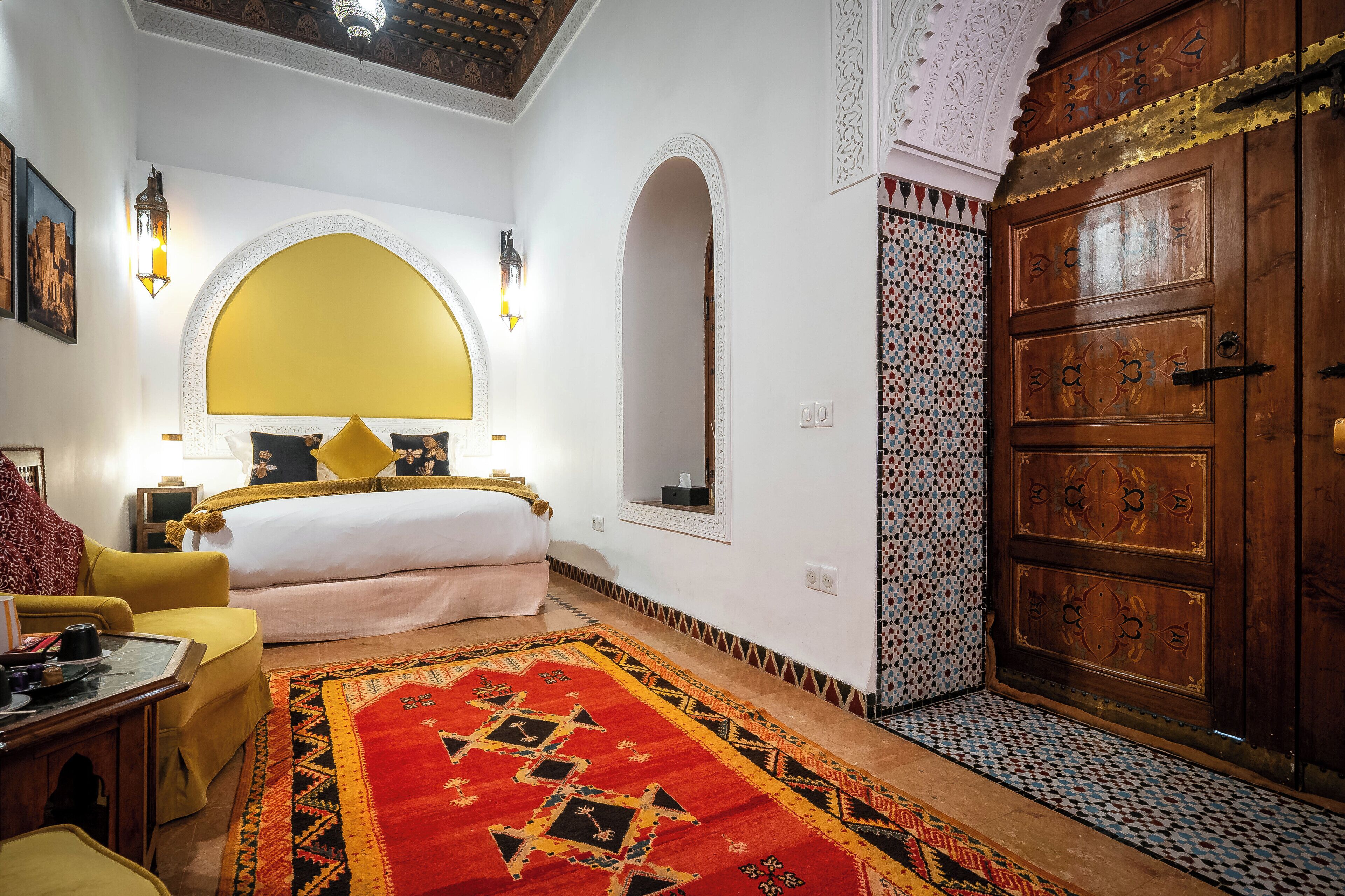 Una habitación con una cama, un sillón amarillo, un tapete de diseño rojo y negro, un armario de madera y un espejo decorativo.