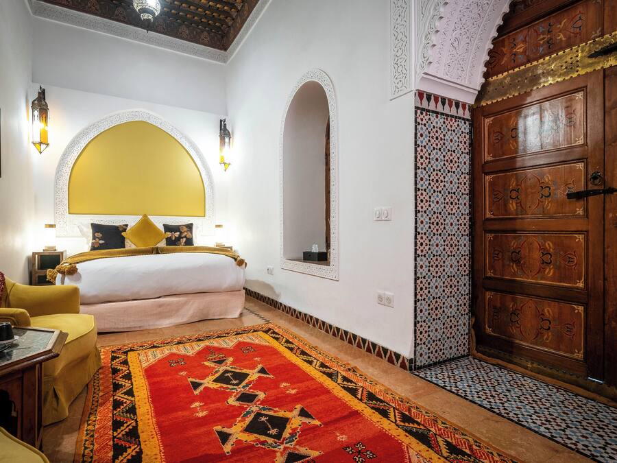 Una habitación con una cama, un sillón amarillo, un tapete de diseño rojo y negro, un armario de madera y un espejo decorativo.