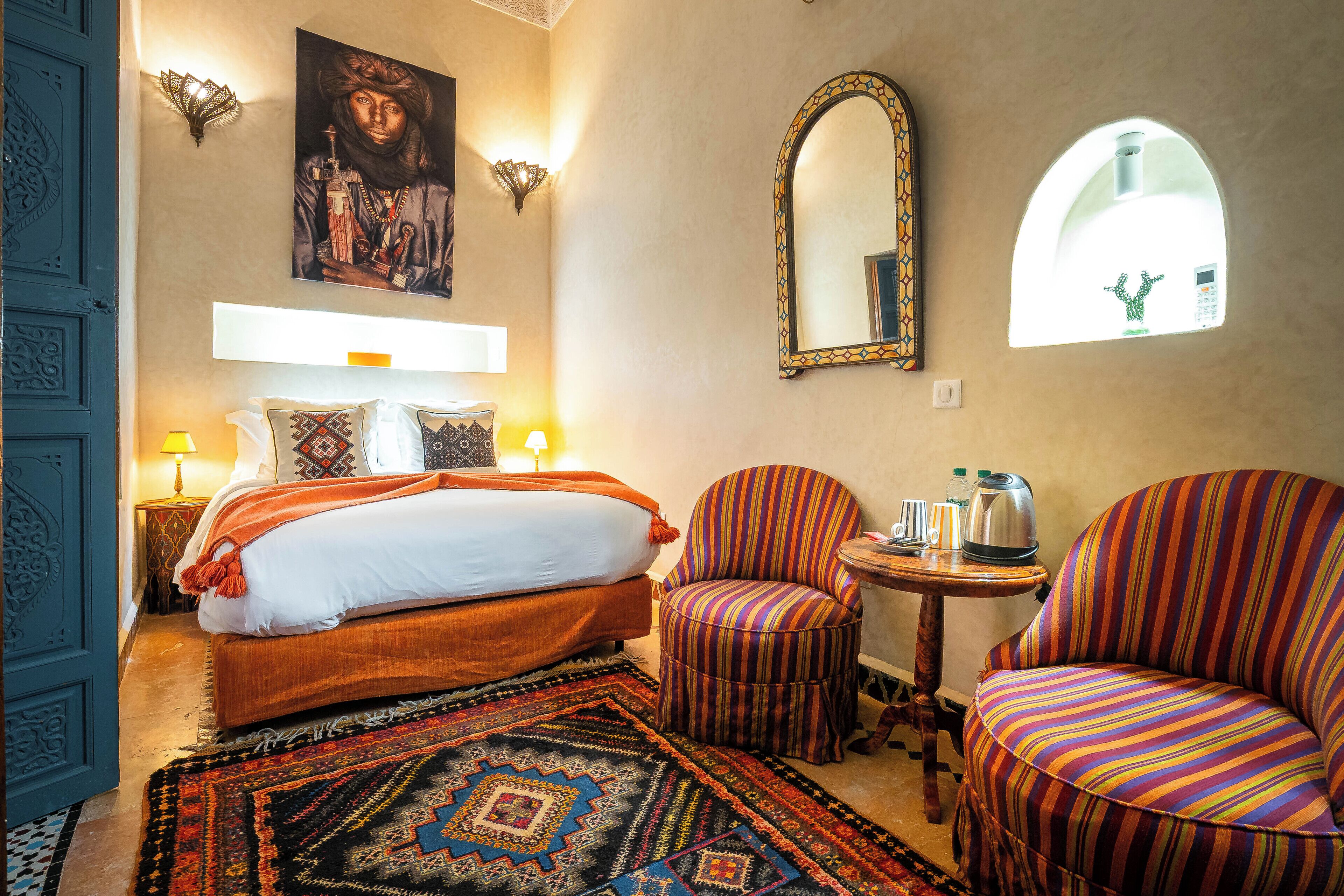 Un dormitorio con cama, una silla, una mesita, un espejo y una ventana.