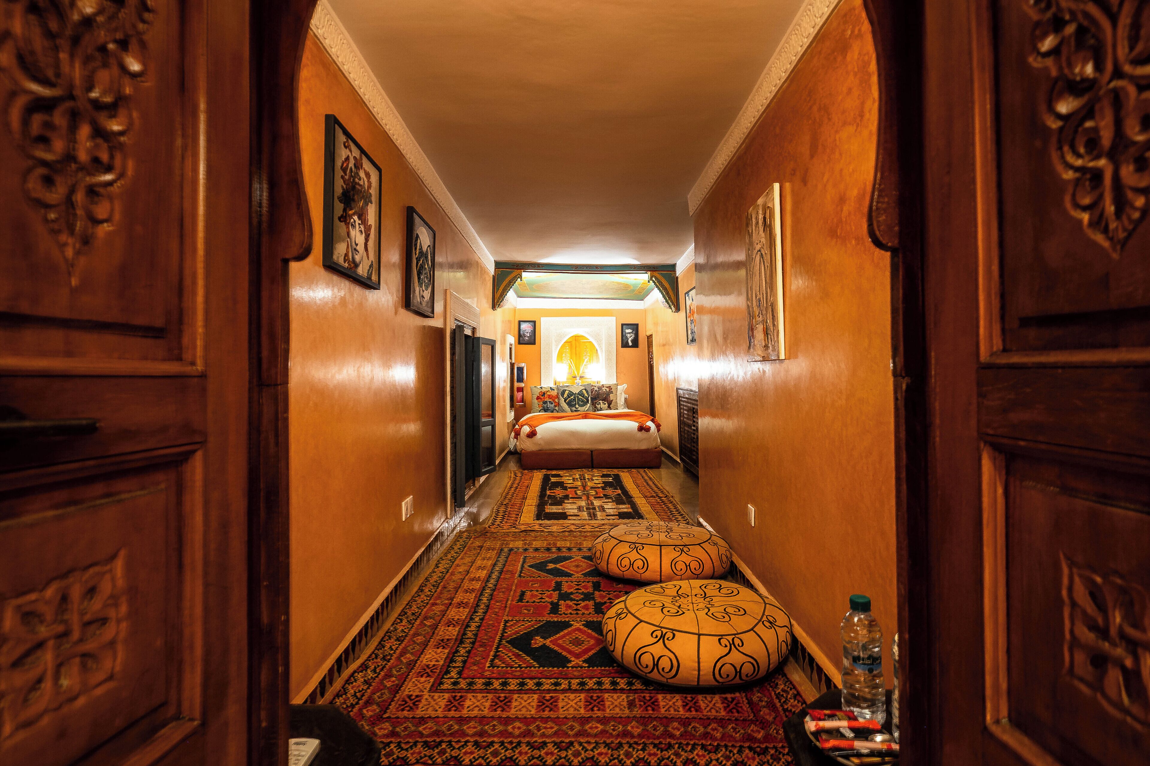A hallway with a patterned carpet, wooden doors, and a bed with pillows and a blanket.