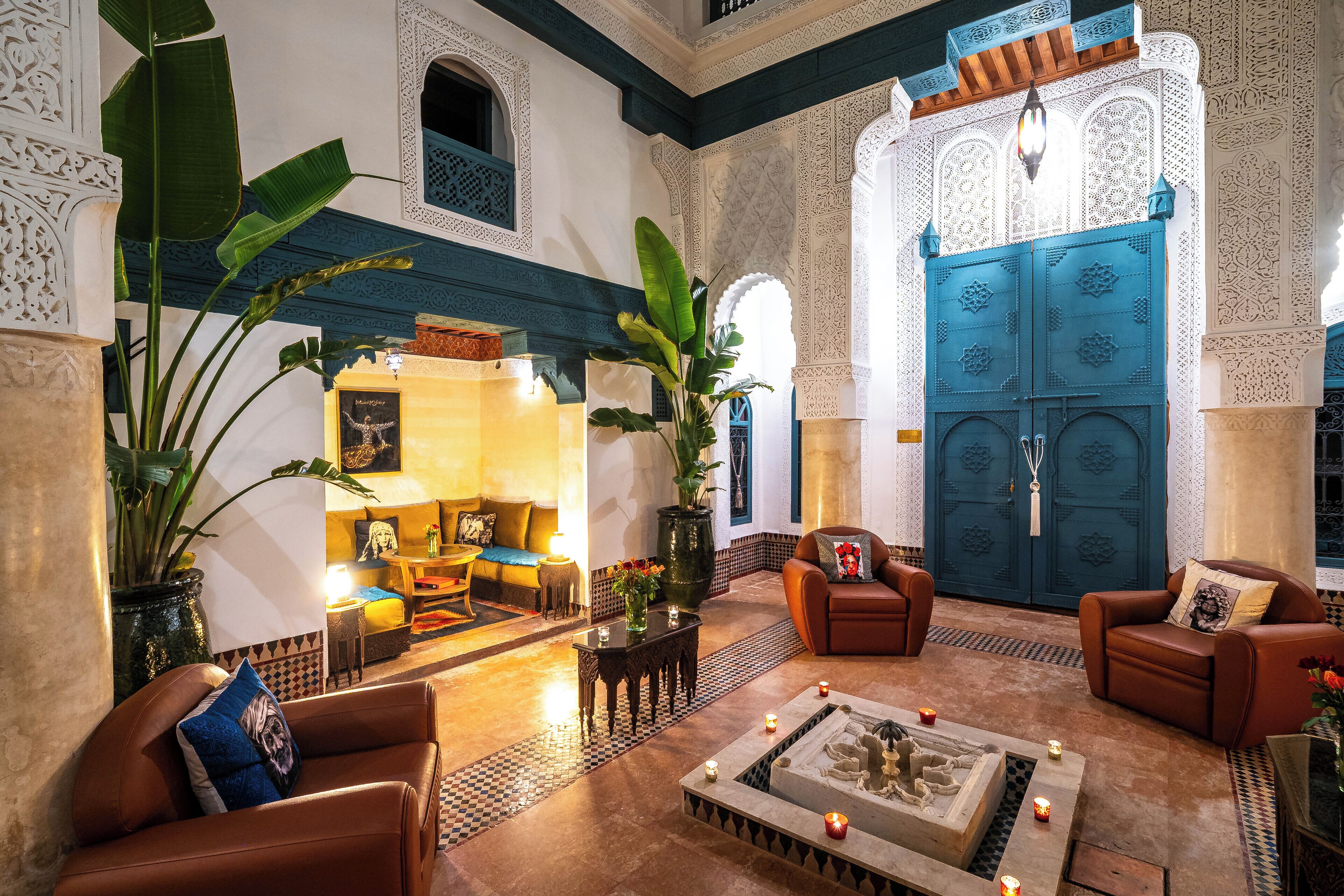 Un interno tradizionale marocchino con un'ampia porta blu, pareti bianche decorate e un'accogliente area salotto con divani in pelle e un tavolino da caffè.