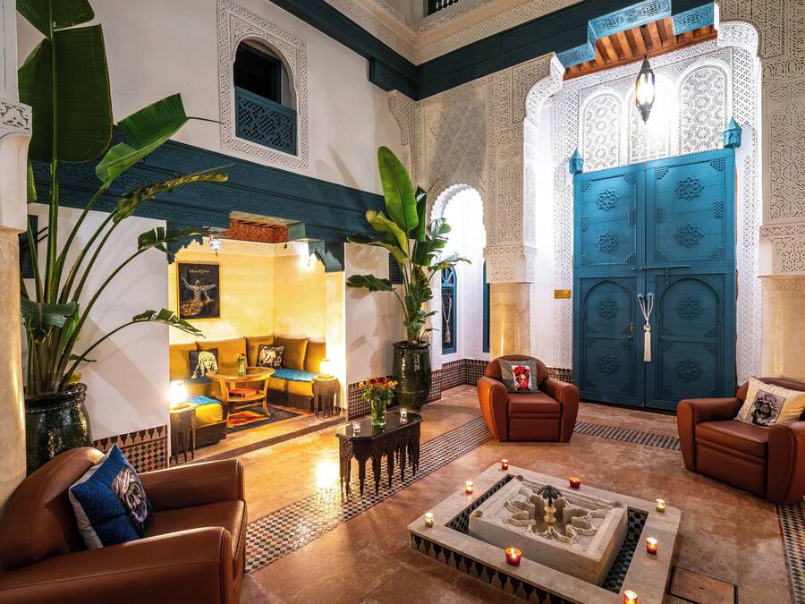 Un interno tradizionale marocchino con un'ampia porta blu, pareti bianche decorate e un'accogliente area salotto con divani in pelle e un tavolino da caffè.