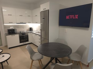 A modern kitchen with white cabinets, a microwave, a coffee maker, and a sink. There is a round table with chairs and a TV mounted on the wall displaying 'NETFLIX'.