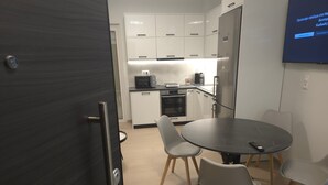 Eine moderne Küche mit weißen Schränken, einer schwarzen Arbeitsplatte und einem Edelstahlkühlschrank. Ein Esstisch mit vier Stühlen und ein an der Wand befestigter Fernseher sind ebenfalls vorhanden.
