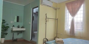A bedroom with a bed, a bedside table, a window with curtains, a bathroom with a sink and mirror, and a potted plant. - Family suite 2 bedrooms for 6 pax (Hà Nội)