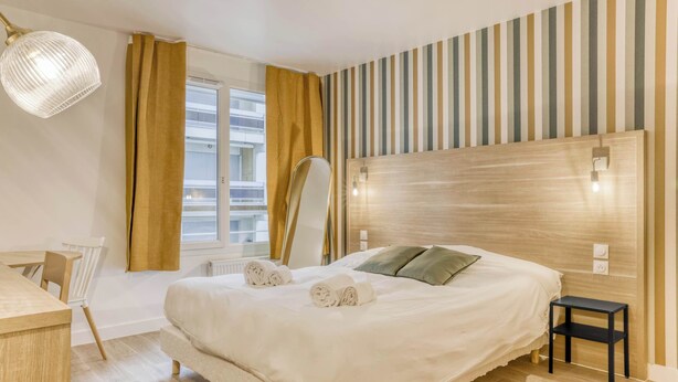 Ein Hotelzimmer mit einem großen Bett, einem Nachttisch, einem Schreibtisch und einem Fenster mit Vorhängen.