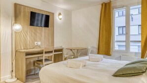Ein Hotelzimmer mit einem Bett, einem Schreibtisch mit Stuhl, einem an der Wand befestigten Fernseher, einem kleinen Tisch mit Stühlen und einem Fenster mit Vorhängen.