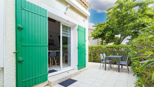 Eine Terrasse mit Tisch und Stühlen, eine Tür mit grünen Fensterläden und ein kleiner Gartenbereich.