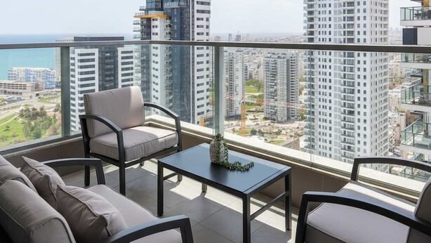 A balcony with a sofa, chairs, and a table, overlooking a cityscape and the sea.