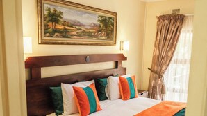Quarto de hotel com uma cama grande, cabeceira de madeira, um quadro acima da cama e uma janela com cortinas.